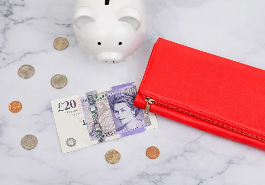 A red zippered wallet with a British £20 note featuring Queen Elizabeth II, surrounded by scattered coins, and a white ceramic piggy bank on a marble surface.